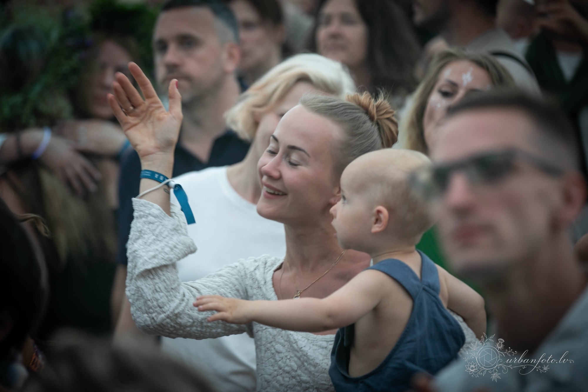 Māte ar mazuli rokās dejo ekstātiskajā dejā BŪTĪBA festivālā 2026, baudot vasaras saulgriežus un Jāņu svinības ģimenēm draudzīgā festivālā Latvijā.