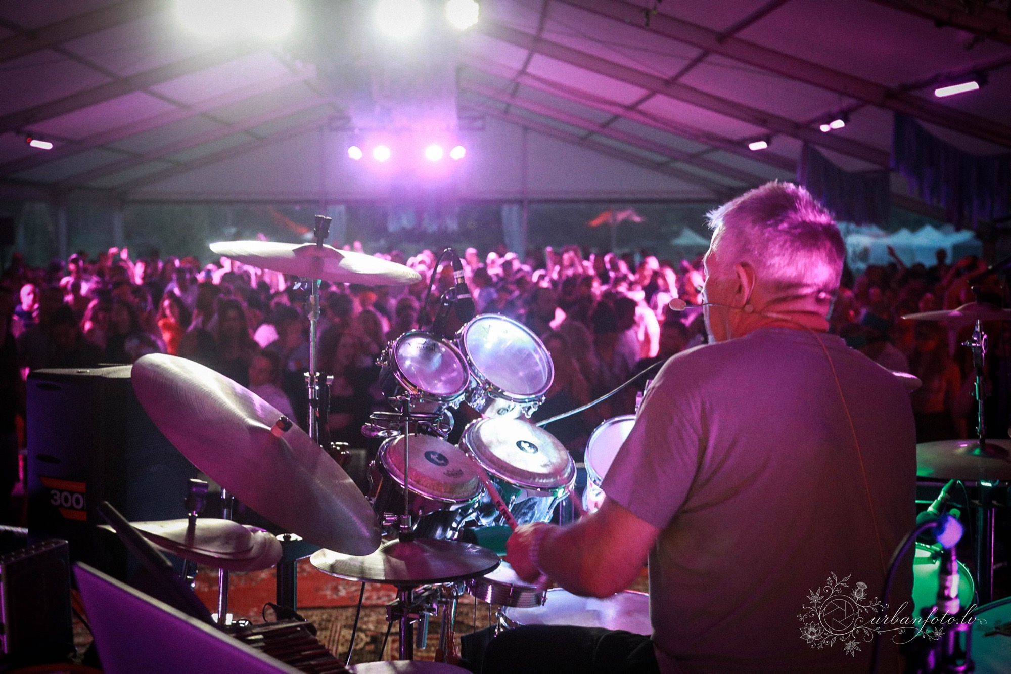 Rishi, seen from behind at a drum and cymbal set, facilitating a trance dance for the crowd under purple stage lights in the tent
