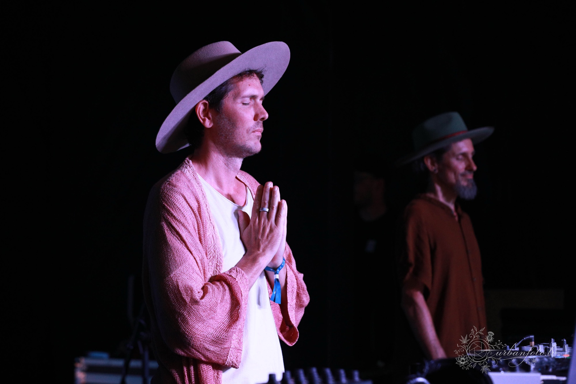 DJ Mose in a wide-brimmed hat and pink cardigan stands with hands in prayer position and eyes closed, preparing for his ecstatic dance set