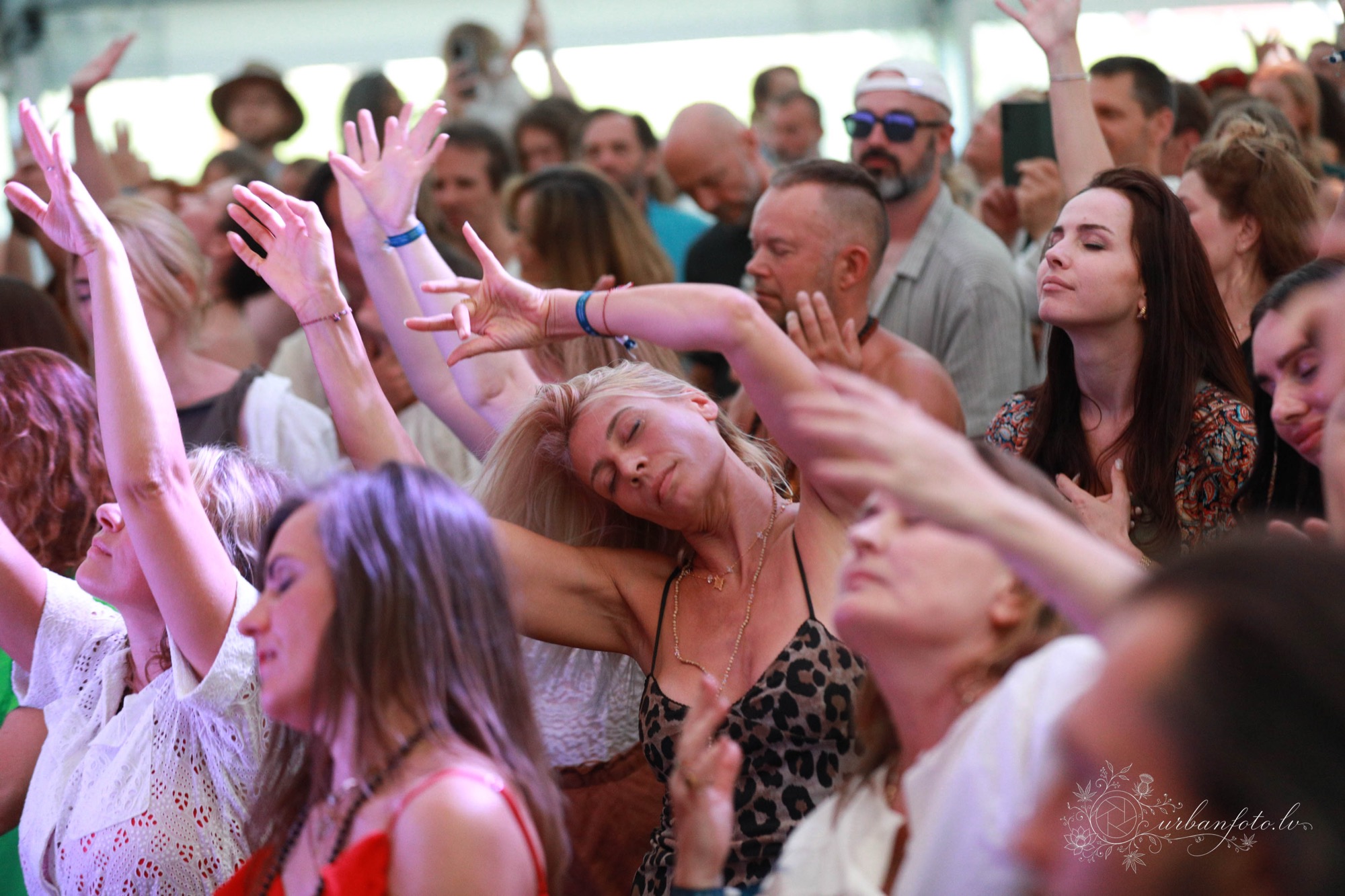 Packed crowd dancing with raised arms and eyes closed, a blonde woman at the center swaying with her head tilted back