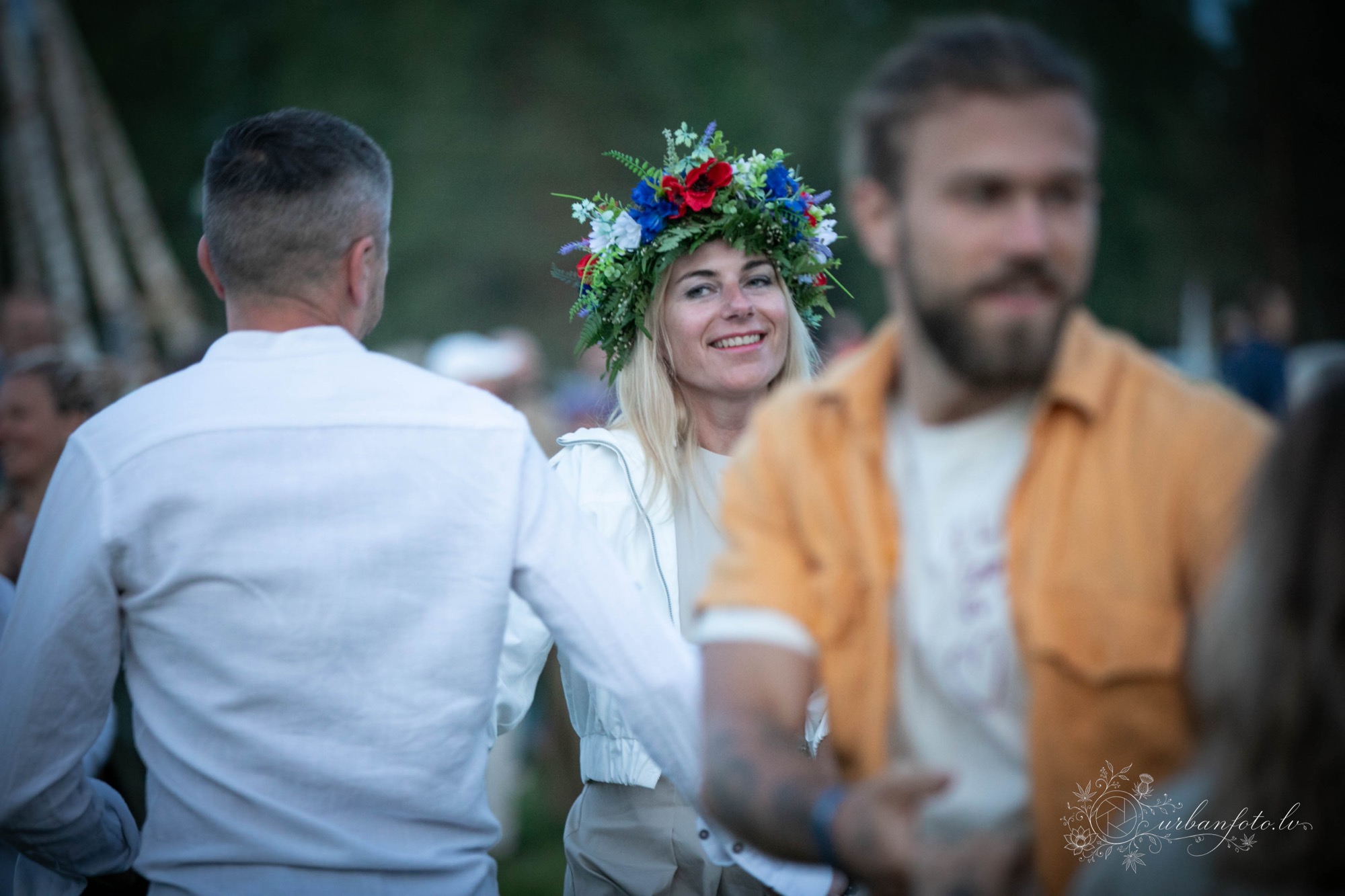 Smaidoša sieviete sarkanu, zilu un baltu ziedu vainagā Rotaļu dejās vakara gaismā