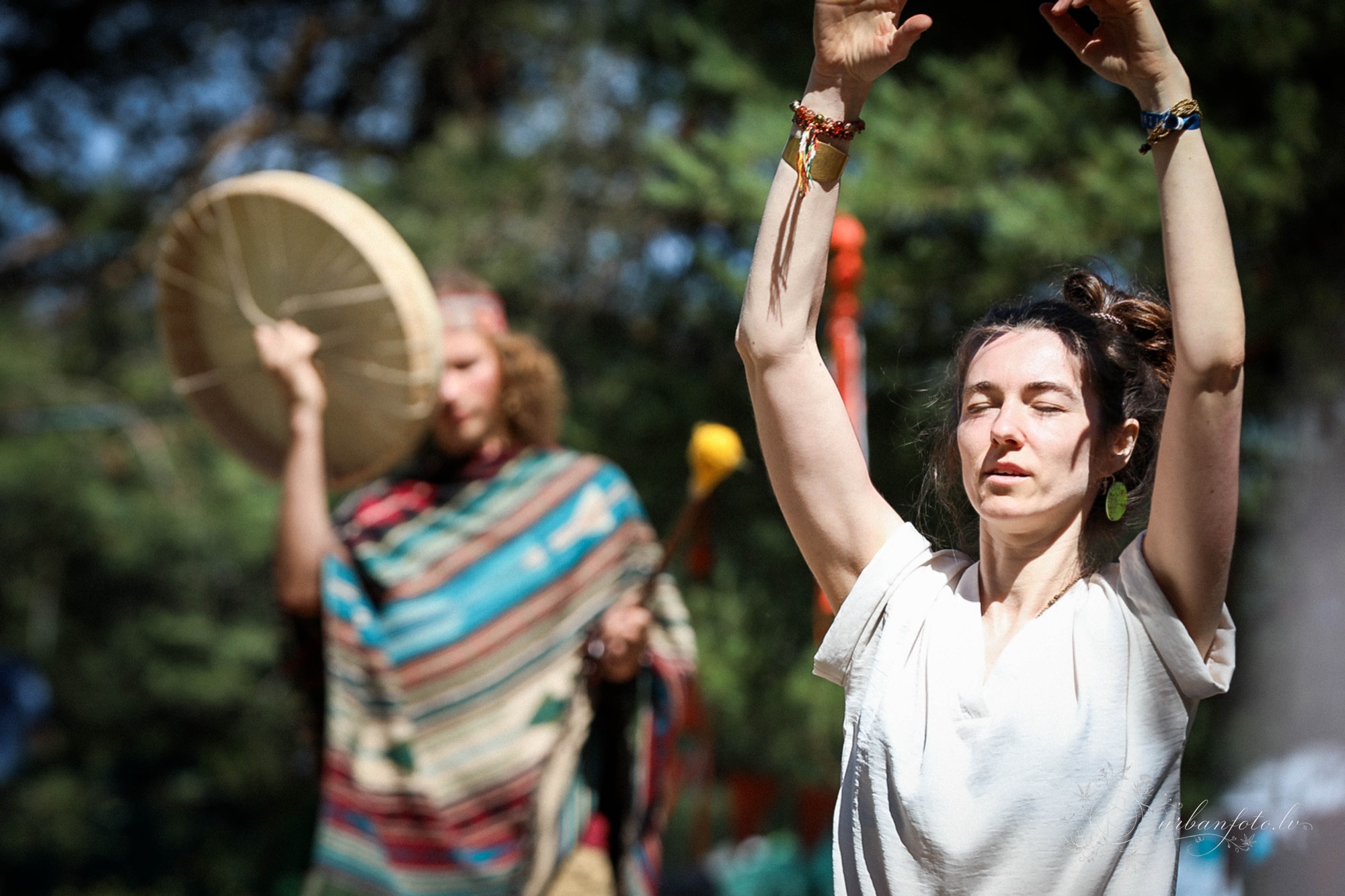 Deep moment of Woman raising her arms during an outdoor ritual accompanied by a participant playing a frame drum at BŪTĪBA Festival 2026