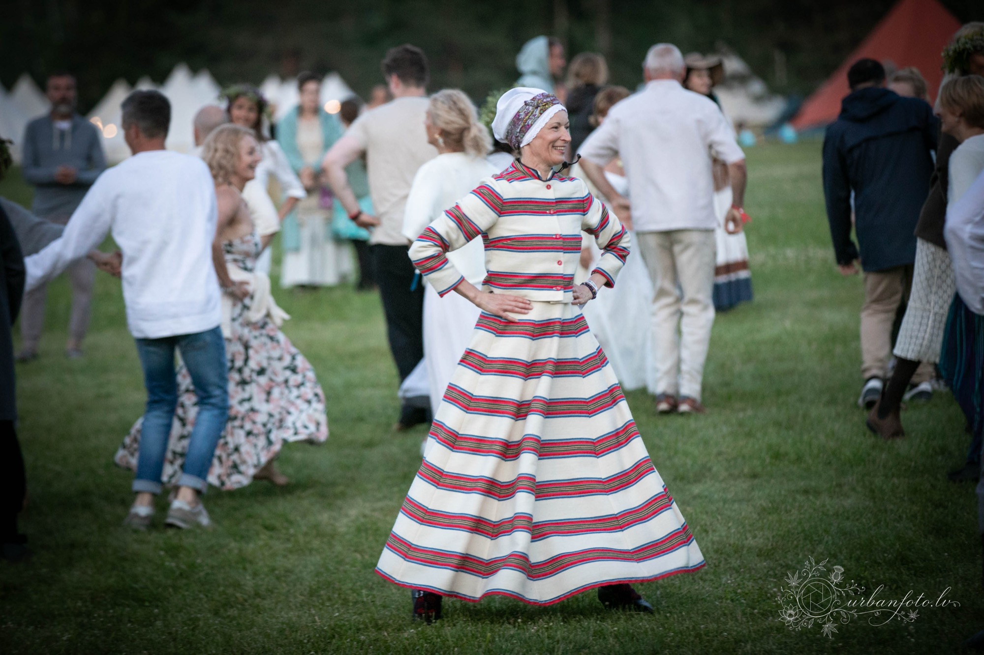 Sandra Bārtniece in a long red and white striped dress and head wrap, hands on hips, with dancers on the grass behind her