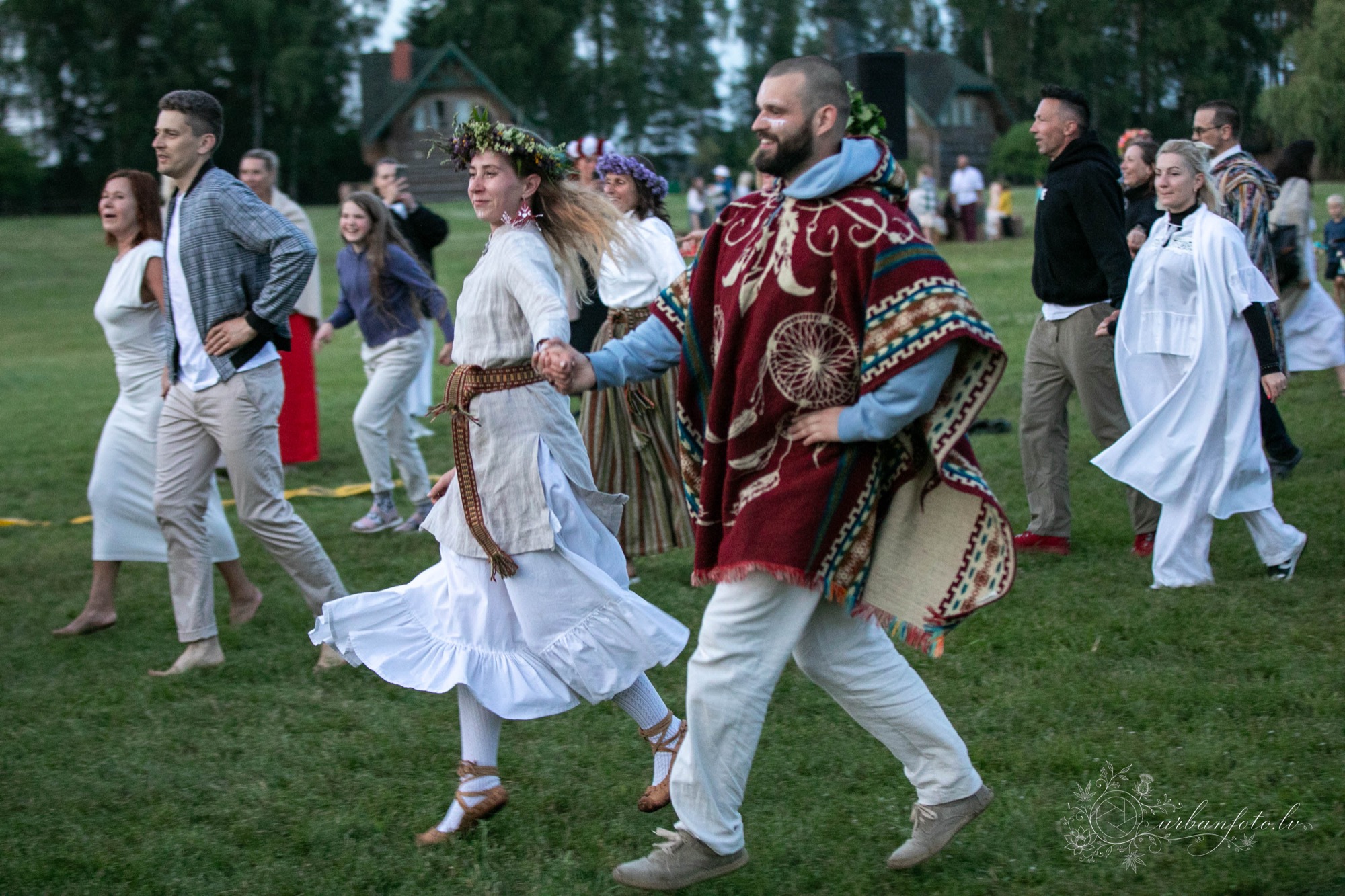 Dalībnieki baltās un tradicionālās drēbēs ar ziedu vainagiem, sadevušies rokās, dejo Rotaļu dejas festivālā Būtība 2026