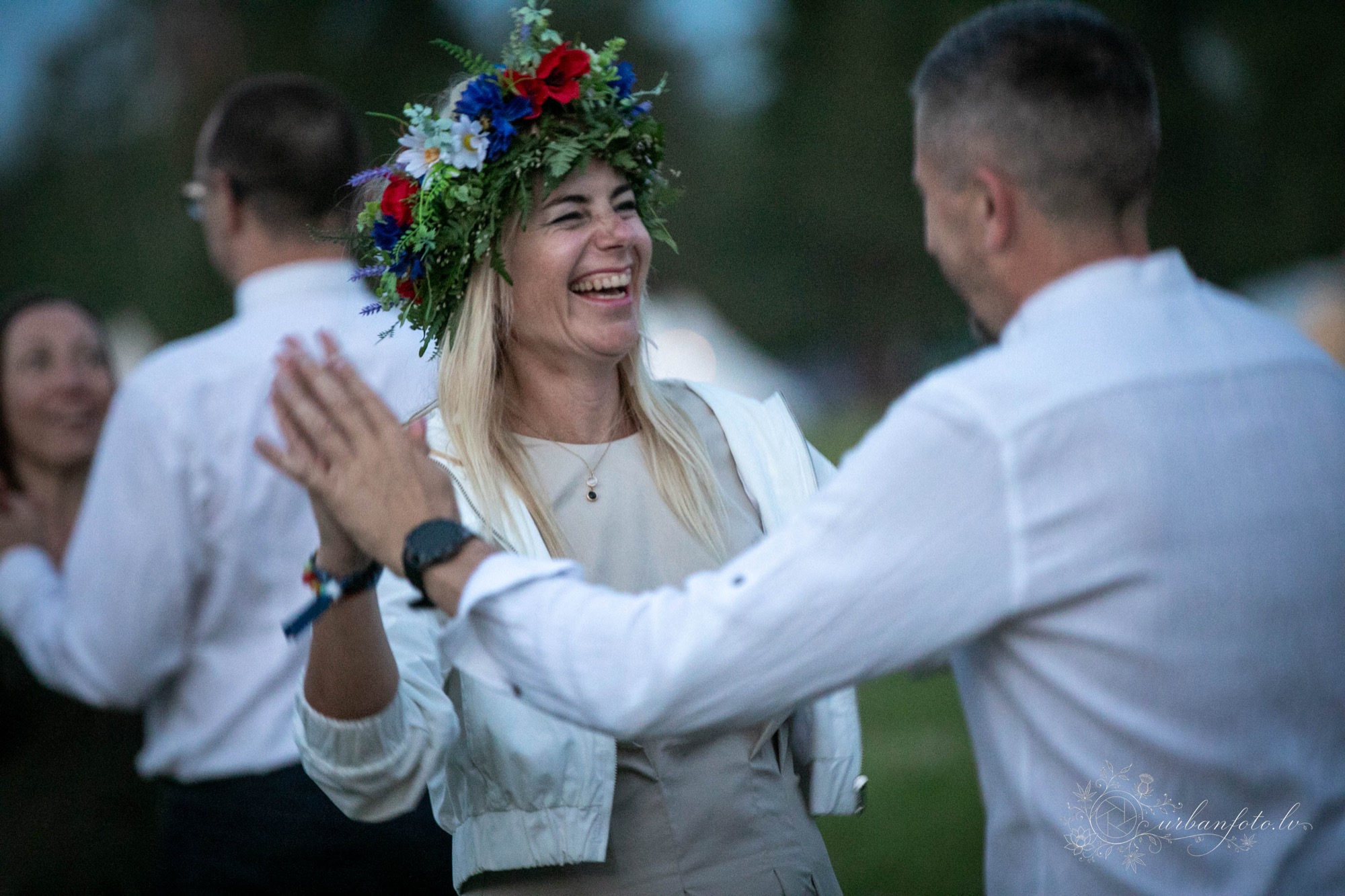 Smejoša sieviete krāsainā pļavas ziedu vainagā sasit plaukstas ar vīrieti baltā kreklā tautu dejās festivālā Būtība 2026