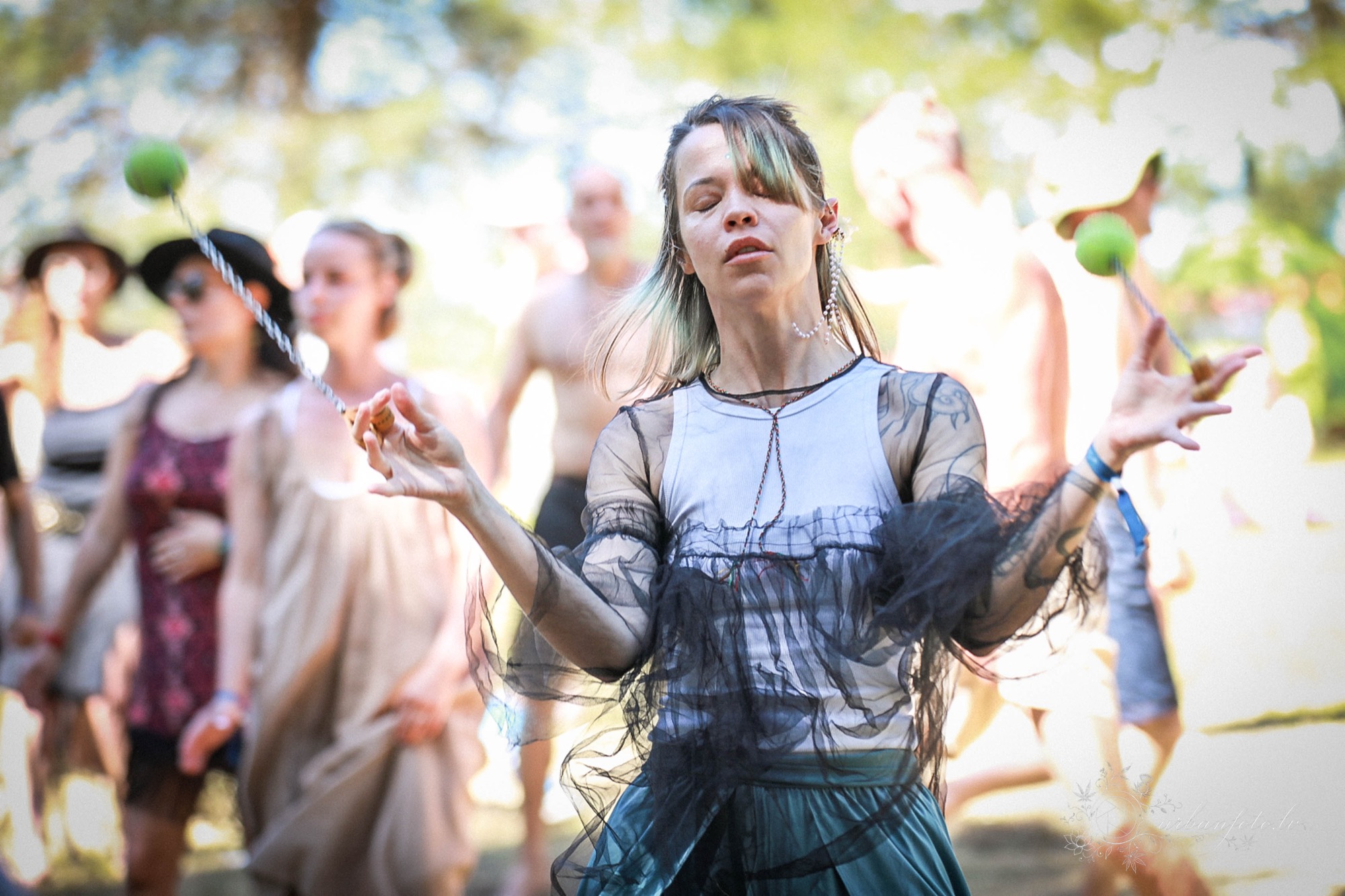 Woman with closed eyes swinging green poi balls on cords while people gather behind her at Būtība Festival 2026