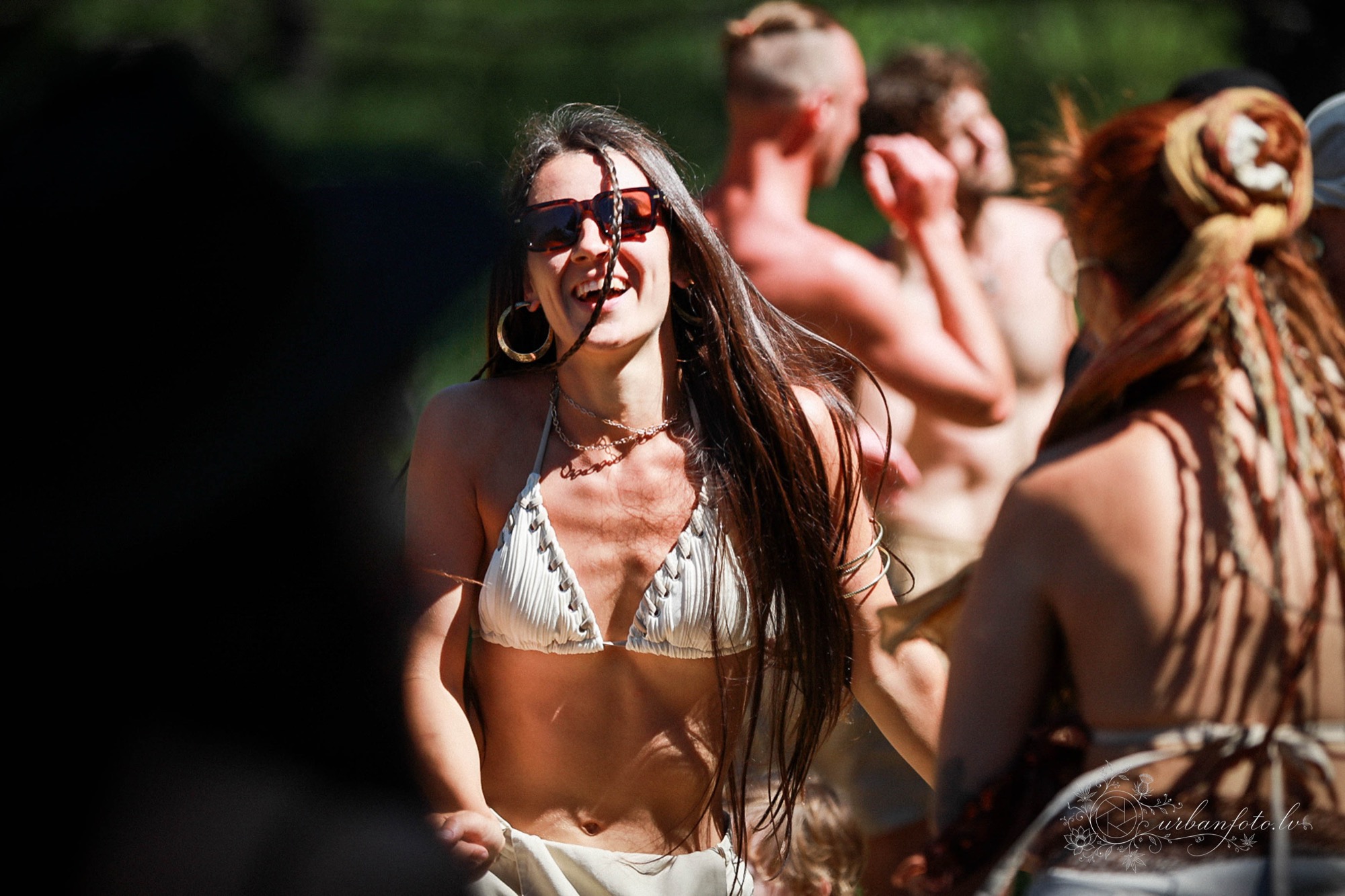Laughing woman with long dark hair and sunglasses dancing in the sun at Būtība Festival 2026