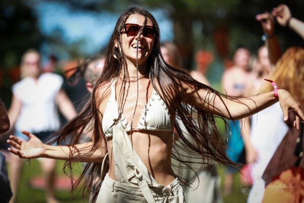 Woman with long dark hair dancing joyfully with open arms in the sunshine at Būtība Festival 2026