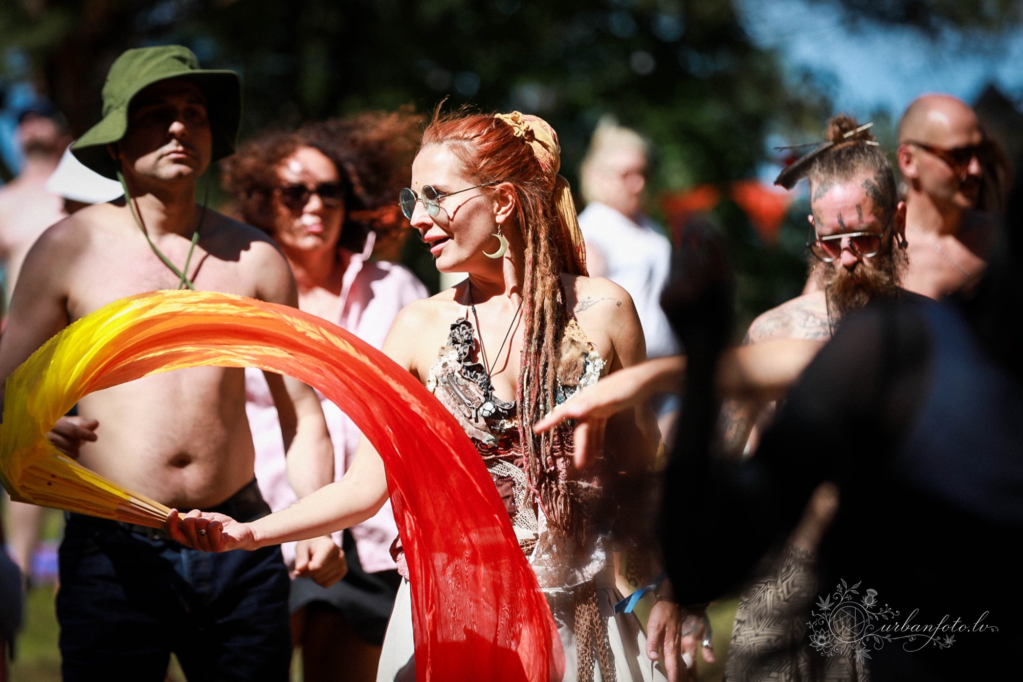 Smaidoša dejotāja virpina oranži sarkanu zīda audumu festivāla dalībnieku vidū festivālā Būtība 2026