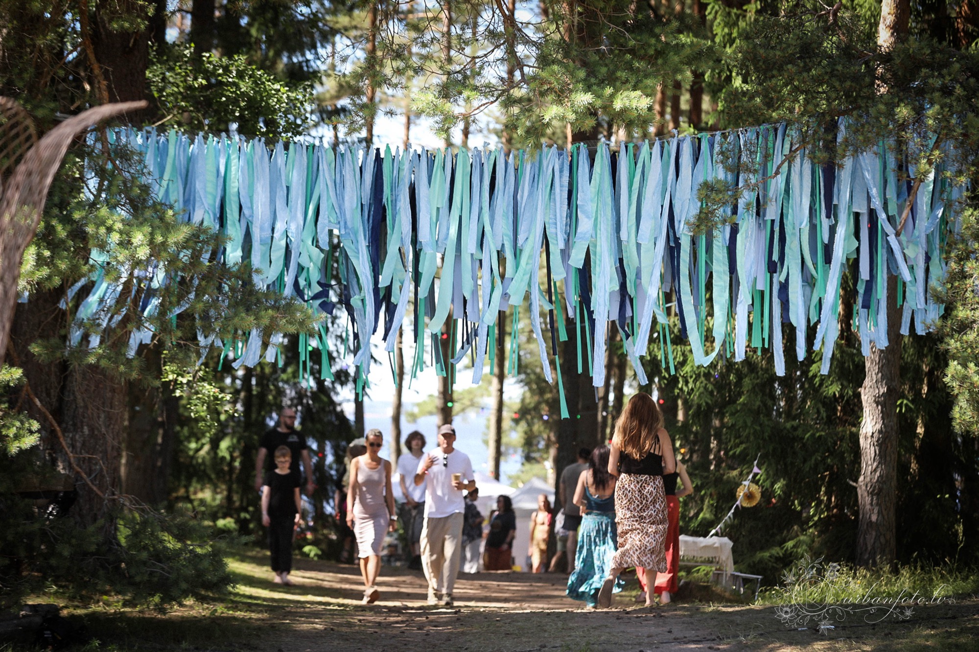 Festival-goers walking along a shaded forest path beneath blue and turquoise fabric decorations at BŪTĪBA Festival 2026