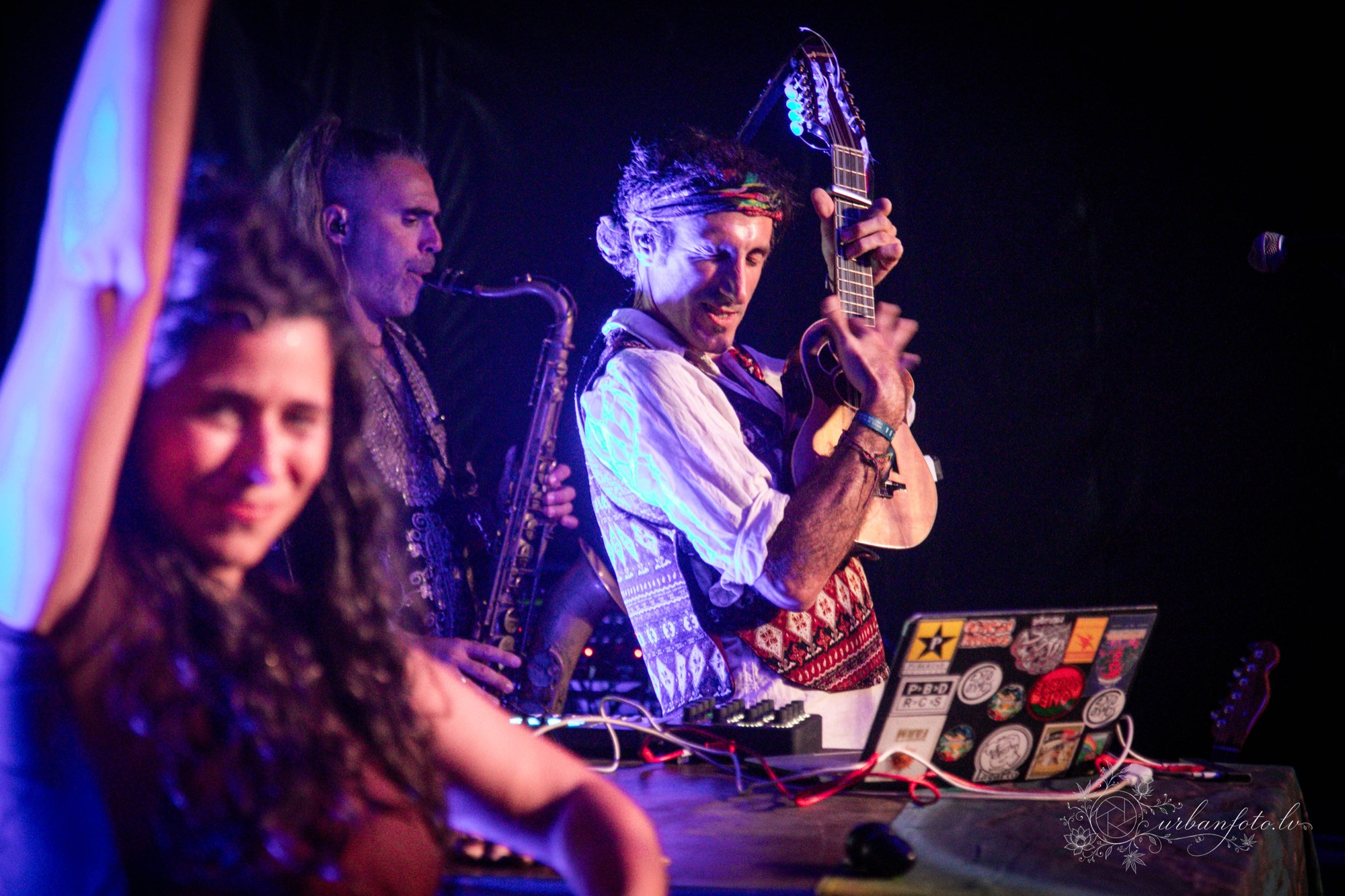 A La Forasteria musician plays an acoustic guitar alongside a saxophonist under the vibrant purple lights of BŪTĪBA Festival 2026. 🎸🎷✨