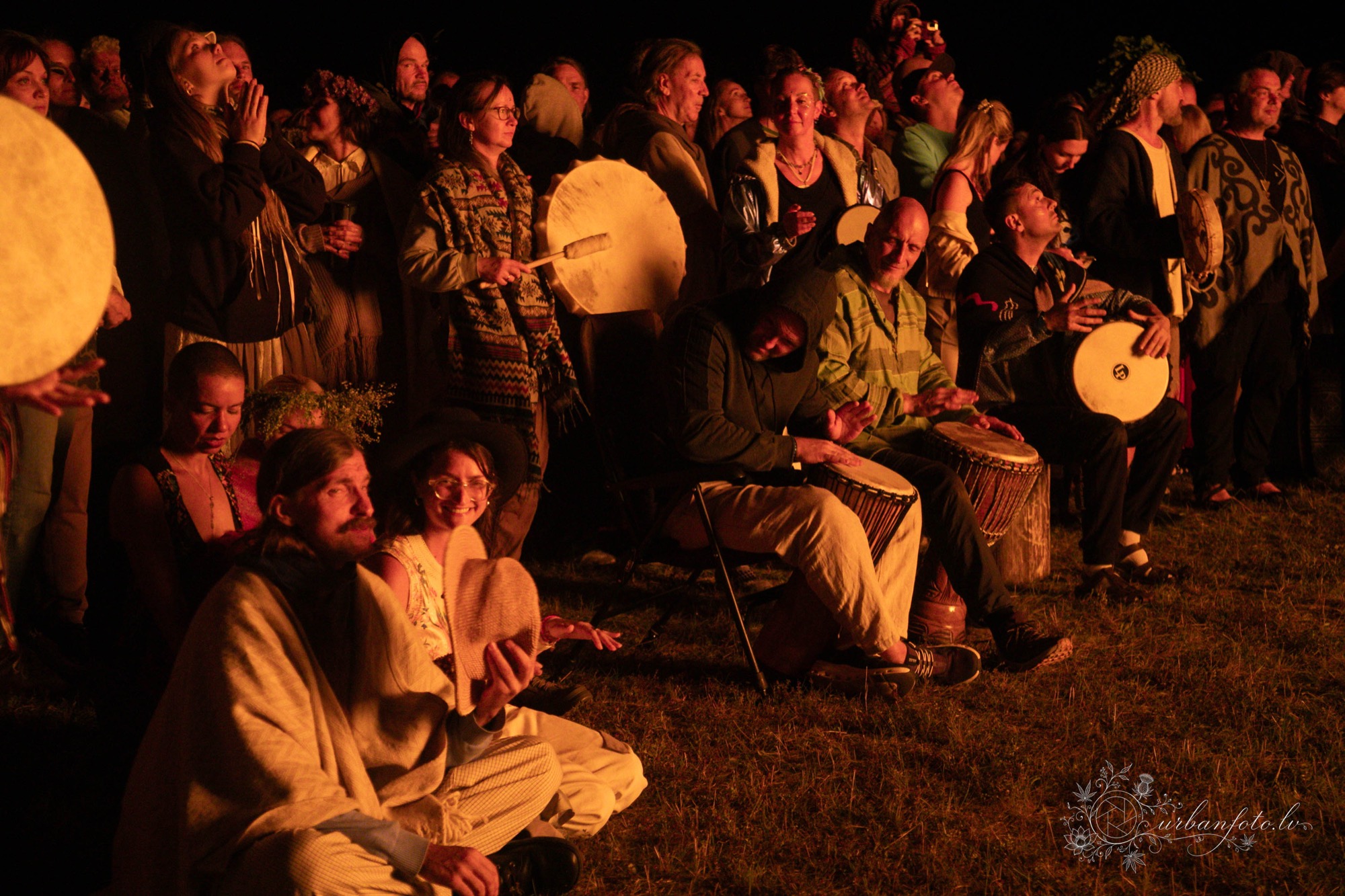 Festival-goers playing hand drums together around a bonfire at BŪTĪBA Festival 2026