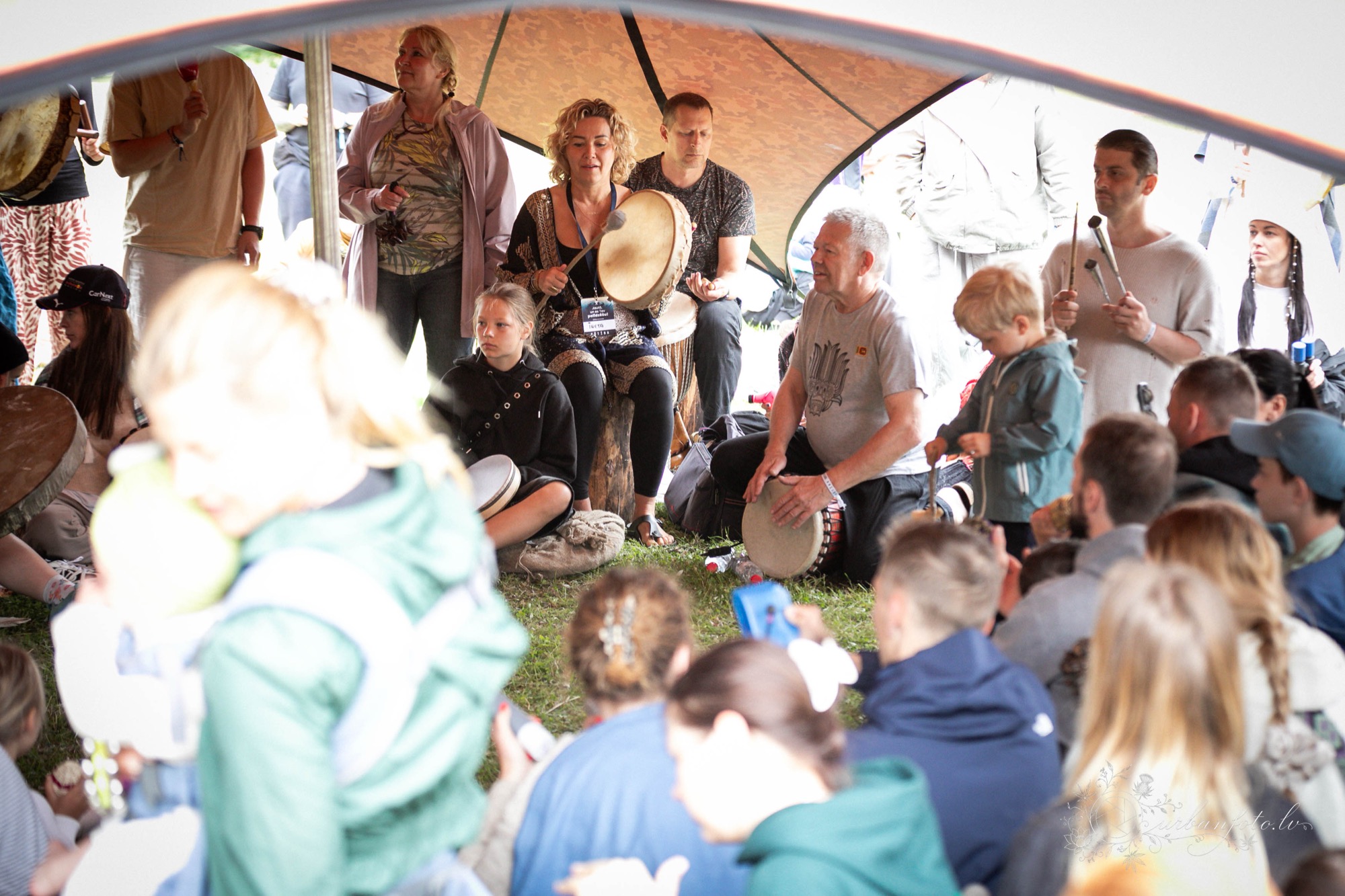 Festival participants of different ages gathered in a circle, playing hand drums and making music together at BŪTĪBA Festival 2026