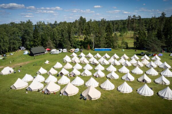 Aerial view of the BŪTĪBA Festival 2026 campsite, with rows of white Glamping tents arranged in a green meadow surrounded by forest