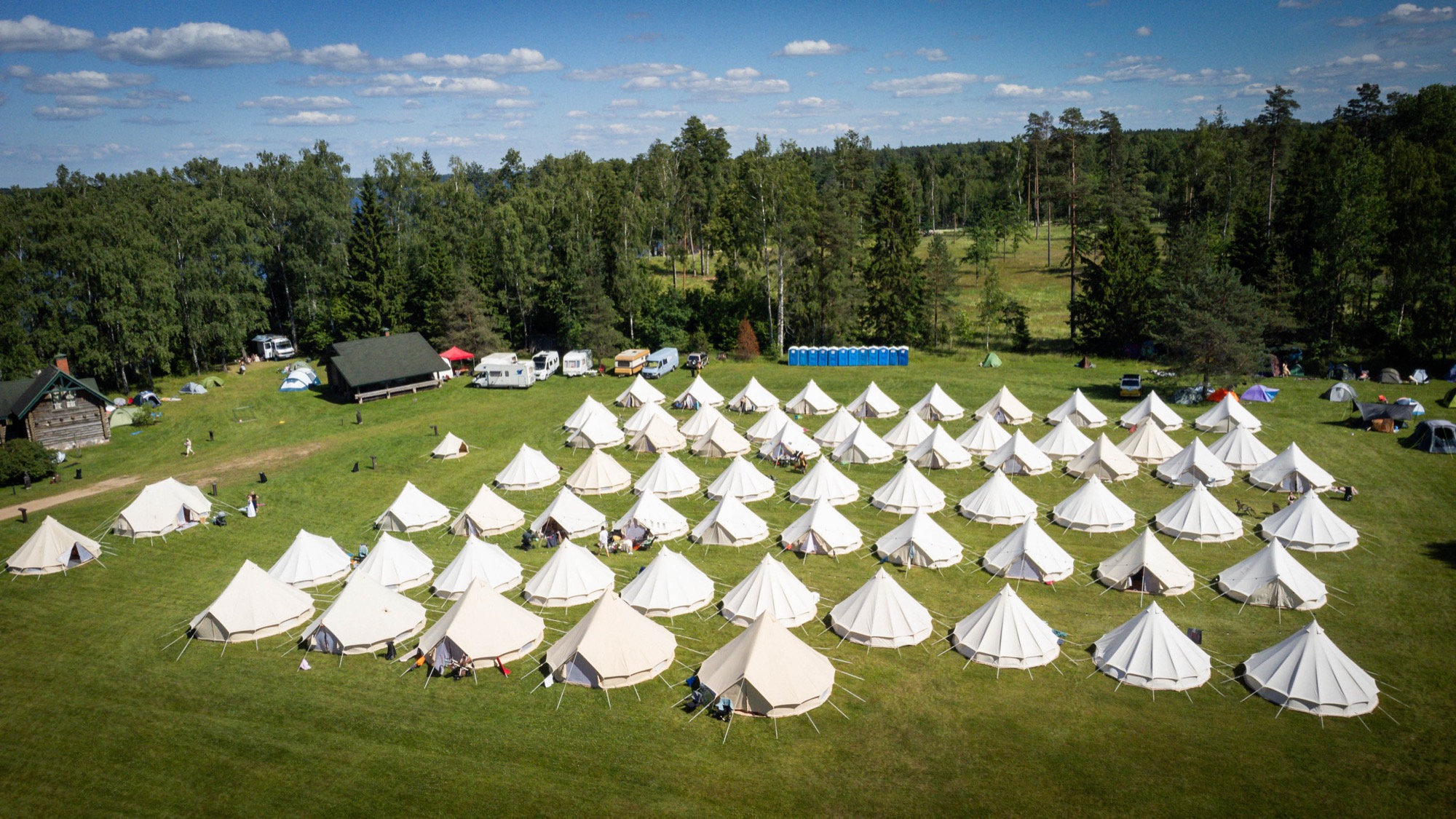 Aerial view of the BŪTĪBA Festival 2026 campsite, with rows of white Glamping tents arranged in a green meadow surrounded by forest