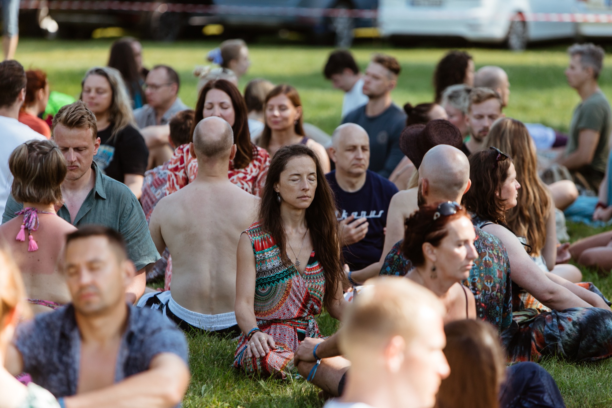 Festival participants sitting outdoors on the grass with their eyes closed during a group meditation at BŪTĪBA Festival 2026