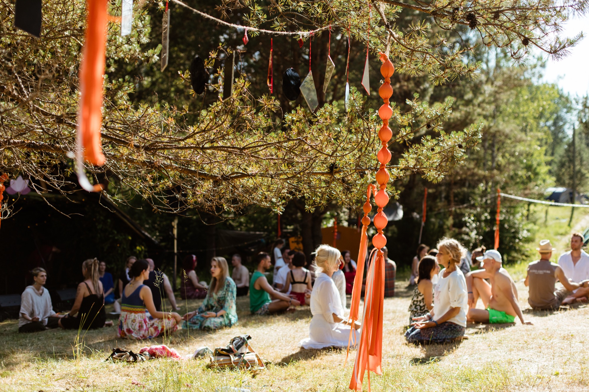 Festival participants sitting together during workshop in a shaded woodland gathering space at BŪTĪBA Festival 2026, surrounded by trees and hanging decorations
