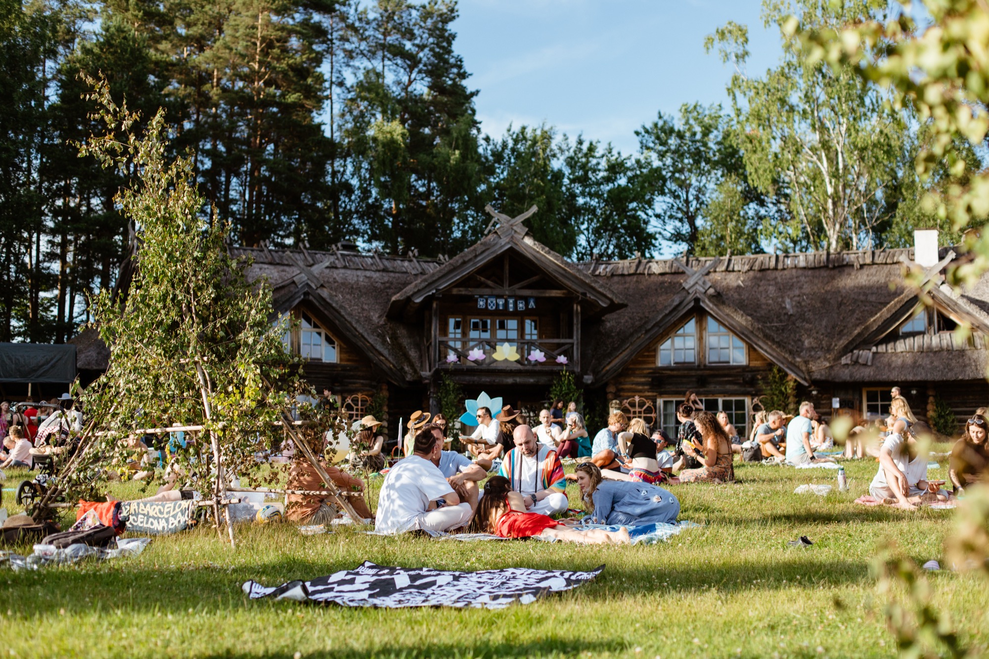 Festival-goers relaxing on the grass outside the traditional wooden BŪTĪBA Festival 2026 venue, surrounded by trees