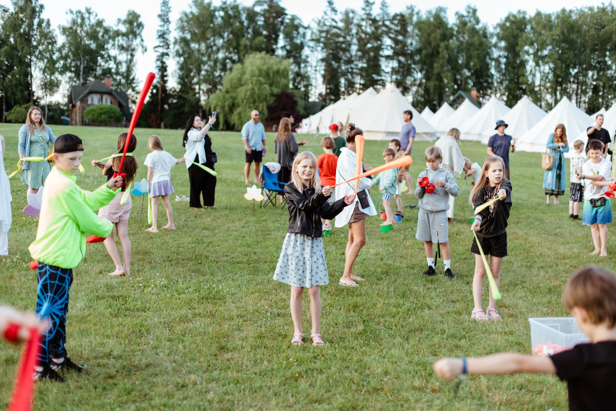 Bērni spēlējas ar krāsainiem poi un piedalās aktivitātēs ģimenēm draudzīgā BŪTĪBA festivālā 2026, svinot vasaras saulgriežus, Jāņus un Līgo Latvijā.