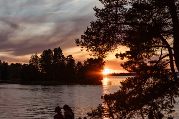 Sunset over the river at BŪTĪBA Festival 2026, with golden light reflecting on the water and pine trees along the shore