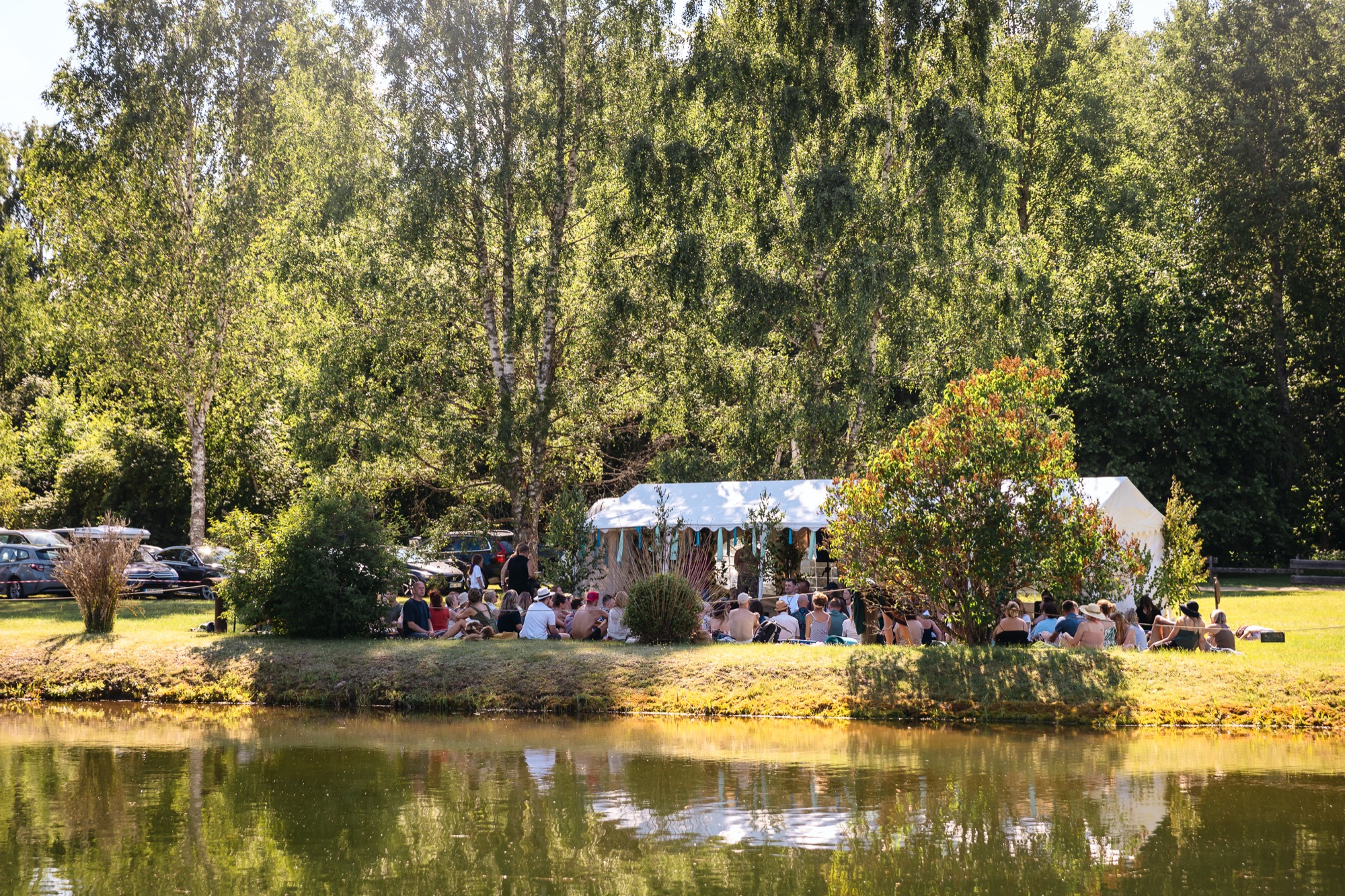 Festival participants gathered for an outdoor workshop beside the water, surrounded by trees at BŪTĪBA Festival 2026