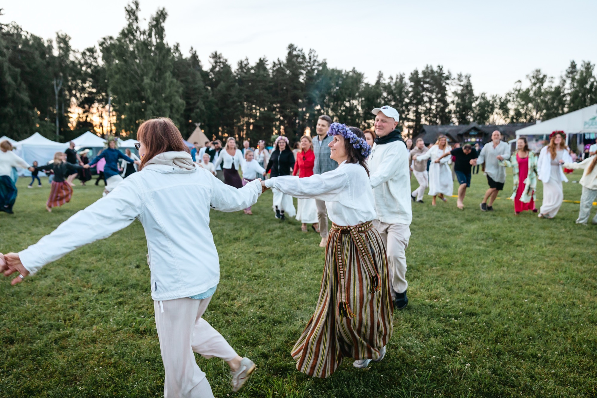 Festival participants dancing latvian folk dances hand in hand in a large circle on the grass at BŪTĪBA Festival 2026 at sunset