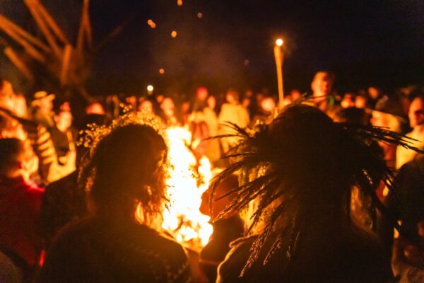Cilvēki ar ziedu vainagiem pulcējas ap ugunskuru, svinot vasaras saulgriežus BŪTĪBA festivālā.