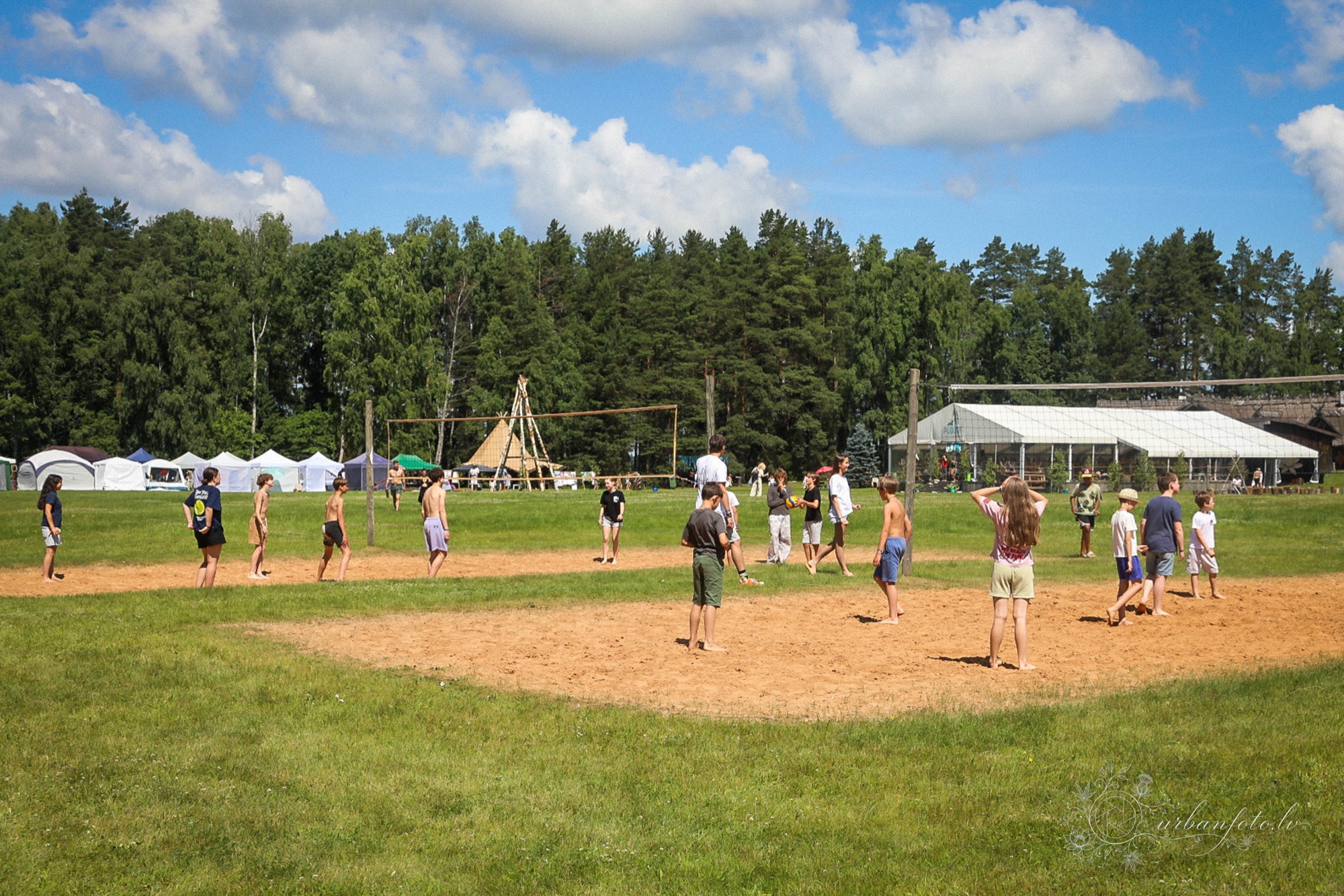 Children playing volleyball on a sandy court surrounded by forest at BŪTĪBA Festival 2026