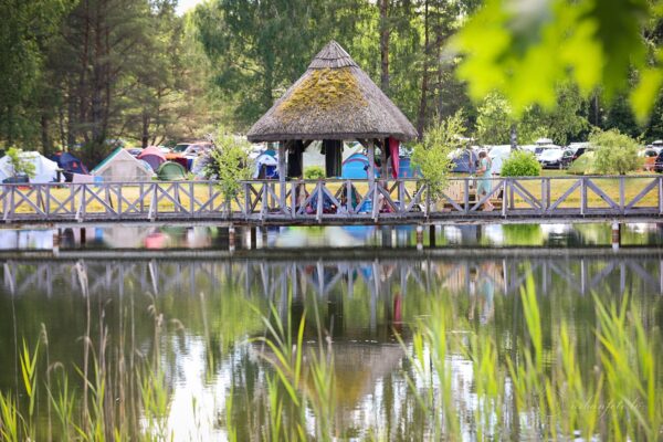 Wooden footbridge crossing a pond beside a thatched shelter and campsite tents at BŪTĪBA Festival 2026