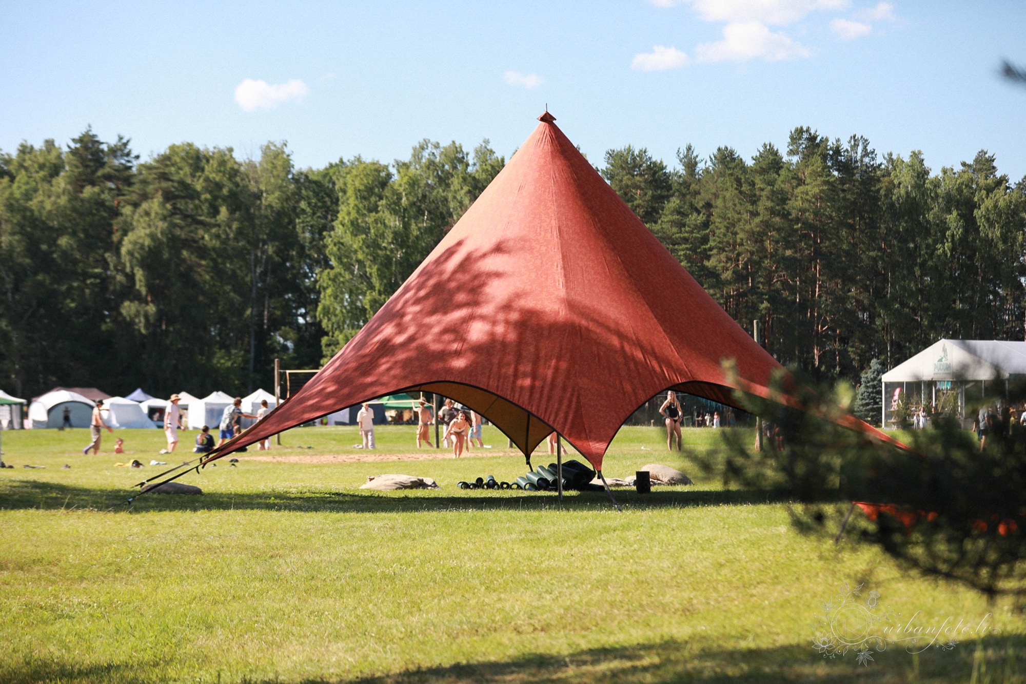 Large red canopy providing shade over a grassy gathering area at BŪTĪBA Festival 2026, surrounded by forest and tents