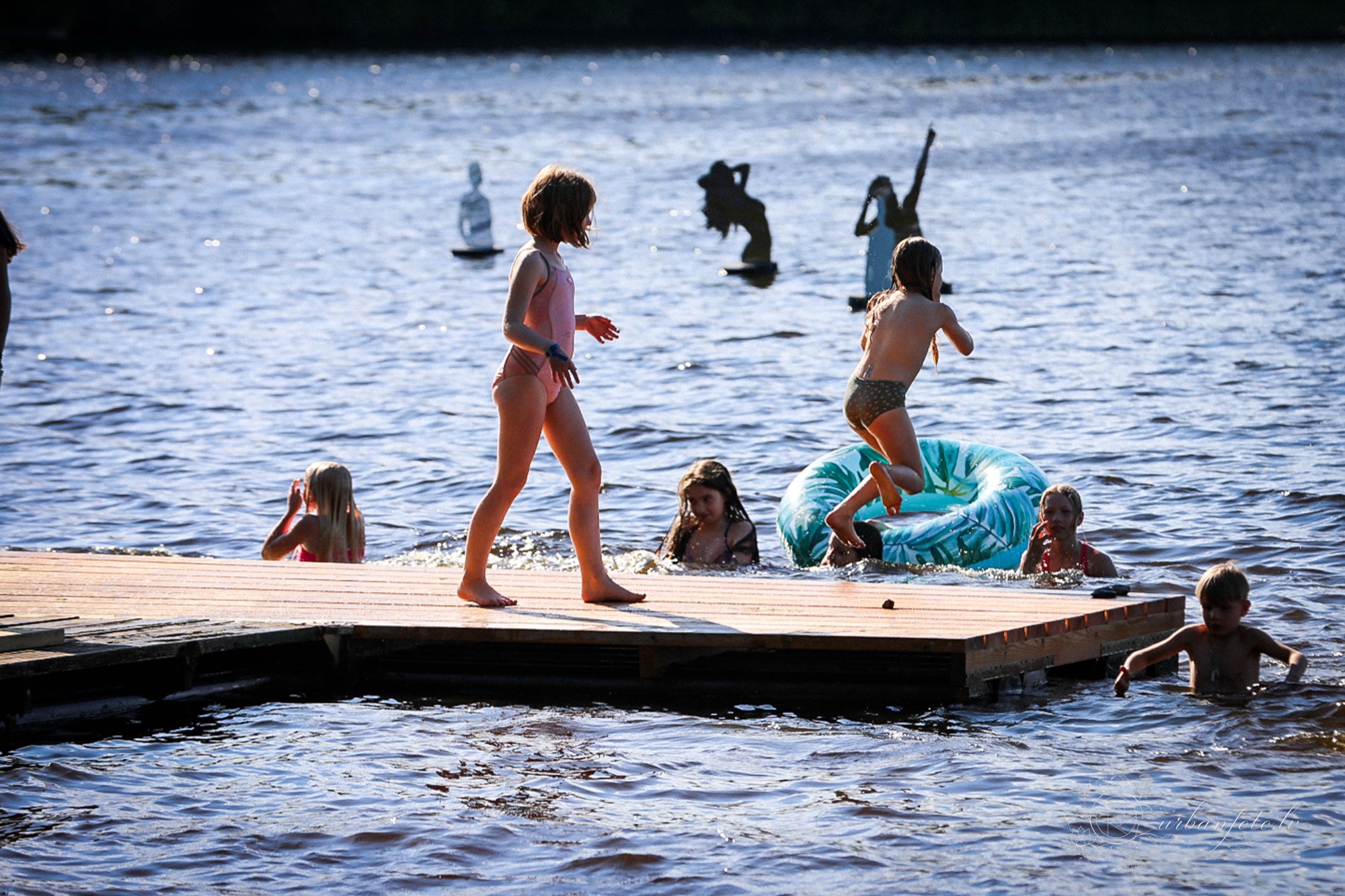 Bērni peldas un rotaļājas Daugavā pie BŪTĪBA festivāla 2026 norises vietas vasaras saulgriežu svinību laikā. Ģimenēm draudzīgs festivāls Latvijā, kur daba un kopā būšana ir svētku daļa.