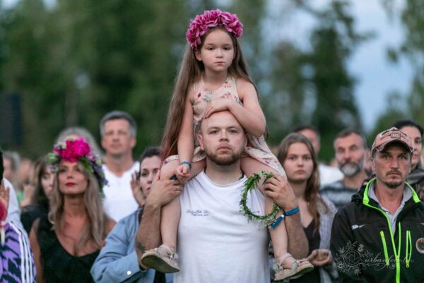 Tētis ar meitu uz pleciem Jāņu svinībās BŪTĪBA festivālā 2026 — ģimenēm draudzīgā vasaras saulgriežu festivālā Latvijā.