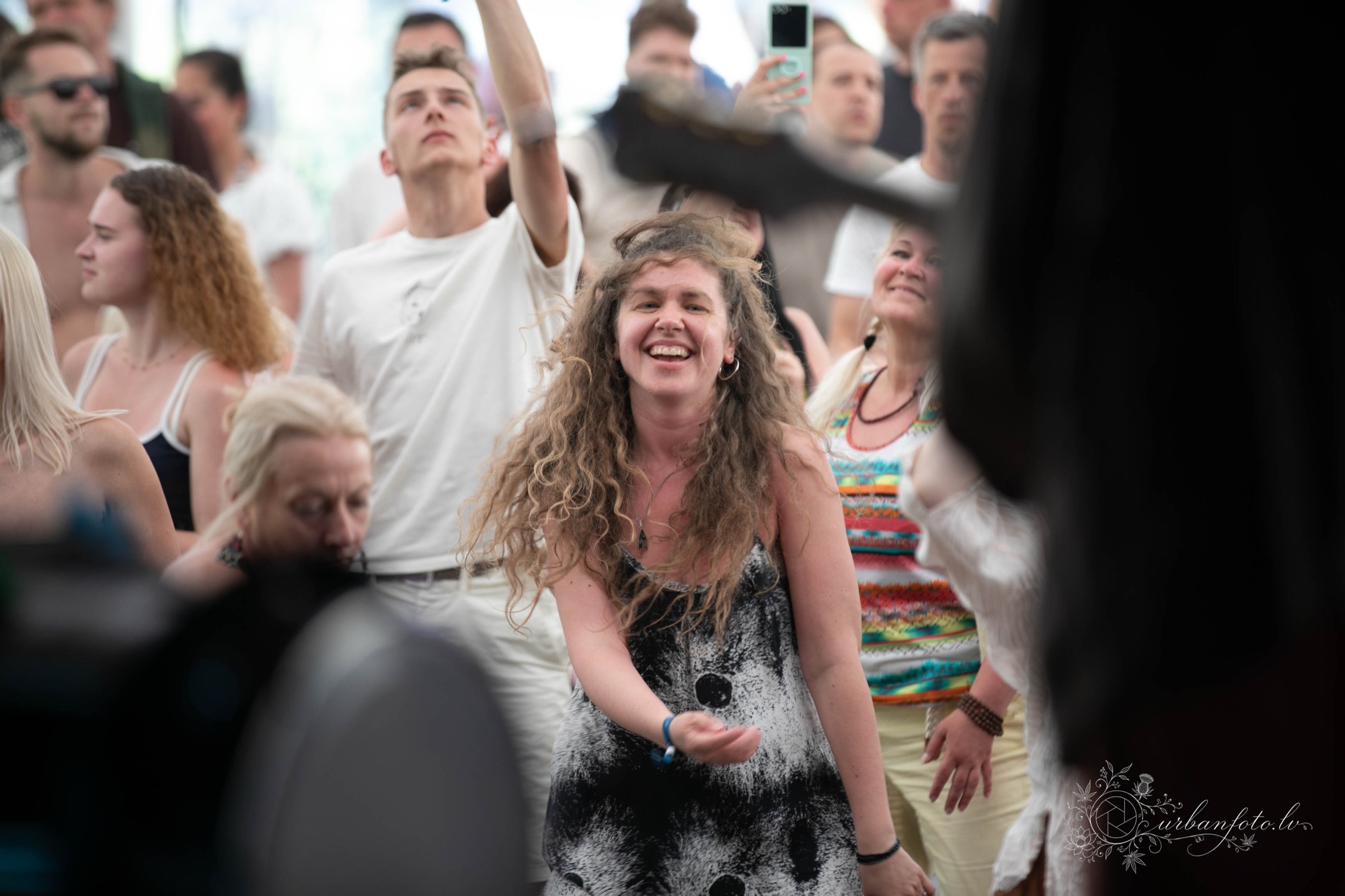 ALT teksts: Sieviete ar cirtainiem matiem priecīgi dejo kopā ar citiem apmeklētājiem BŪTĪBA festivāla vasaras saulgriežu svinībās.