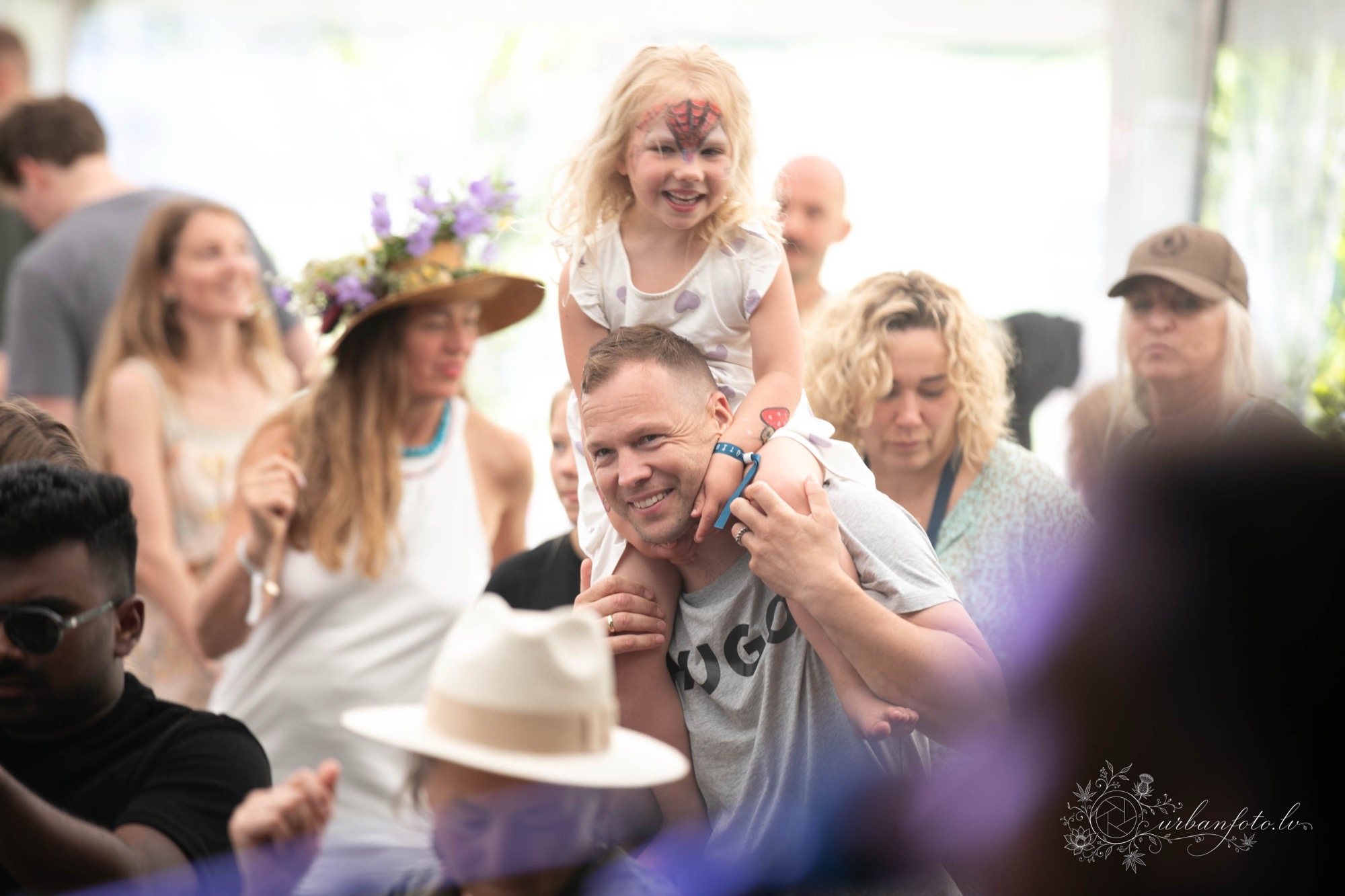 Tētis ar meitu uz pleciem BŪTĪBA festivāla 2026 ģimenēm draudzīgajā vasaras saulgriežu un Jāņu svinību atmosfērā.