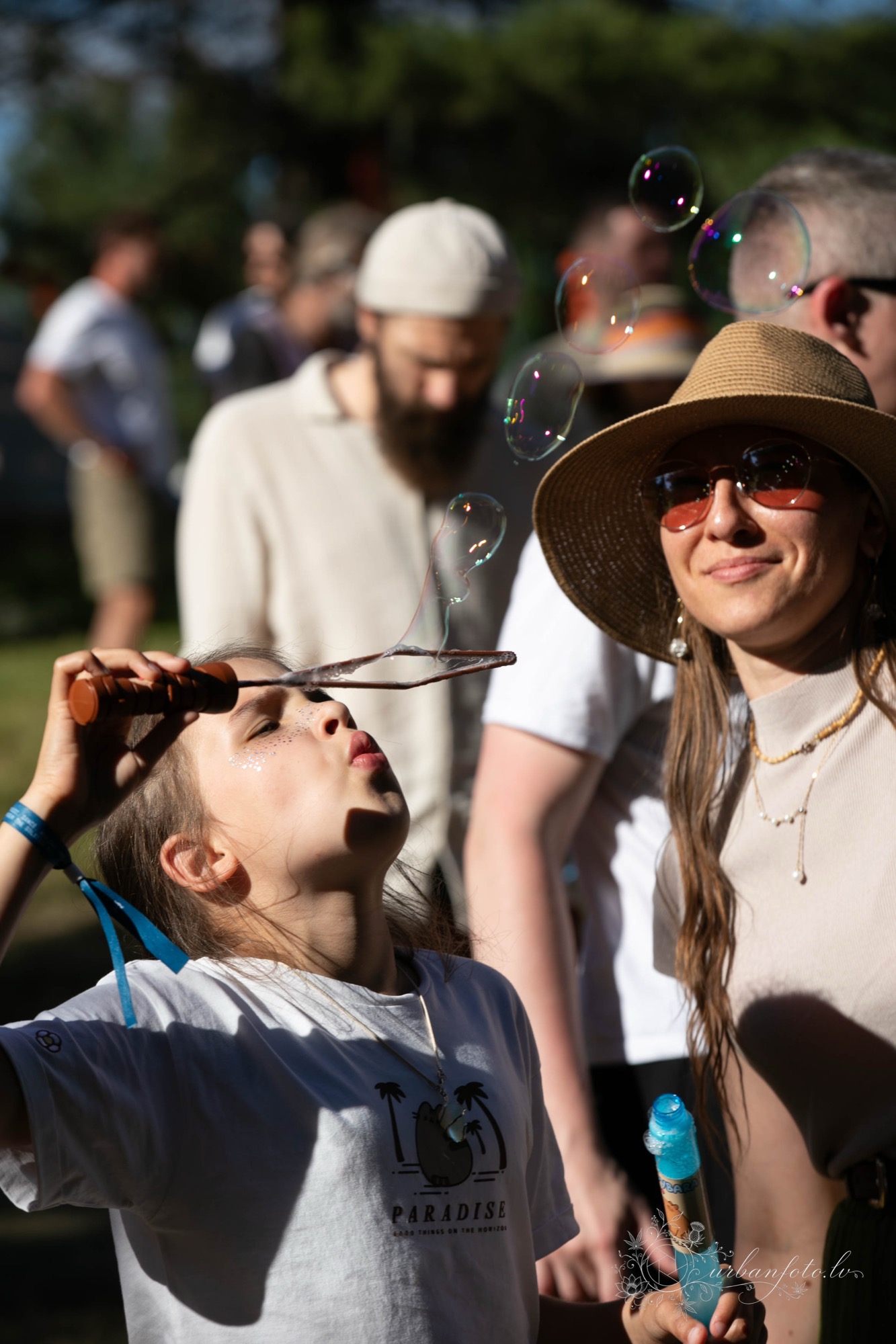 Bērns pūš ziepju burbuļus BŪTĪBA festivāla laikā, kamēr blakus sieviete bauda saulaino dienu.